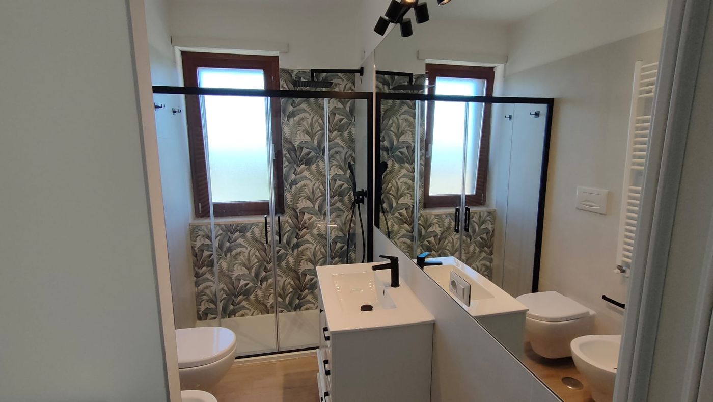 Bagno ristrutturato con parete doccia a foglie verdi, box doccia nero e mobile bianco